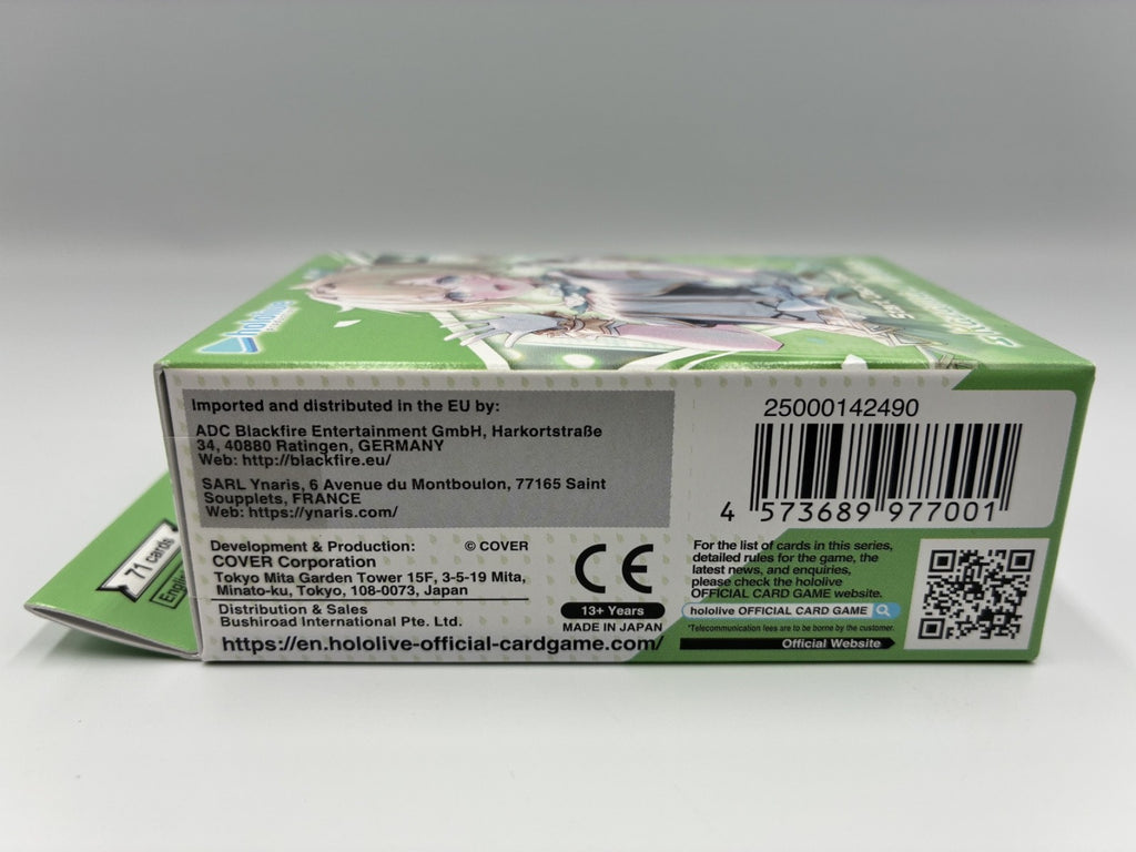 The back of a green and white card game box, showing printed text, a barcode, a QR code, and character artwork, photographed against a light gray background.