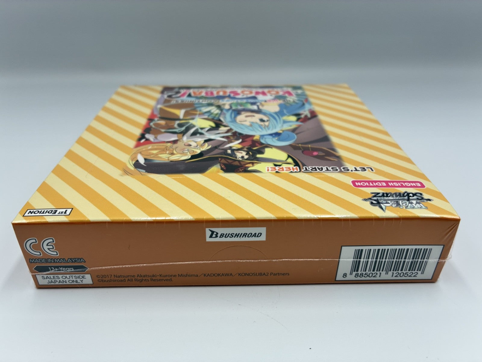Trading card game box featuring a yellow and orange striped design with character artwork on top, photographed on a neutral gray background.