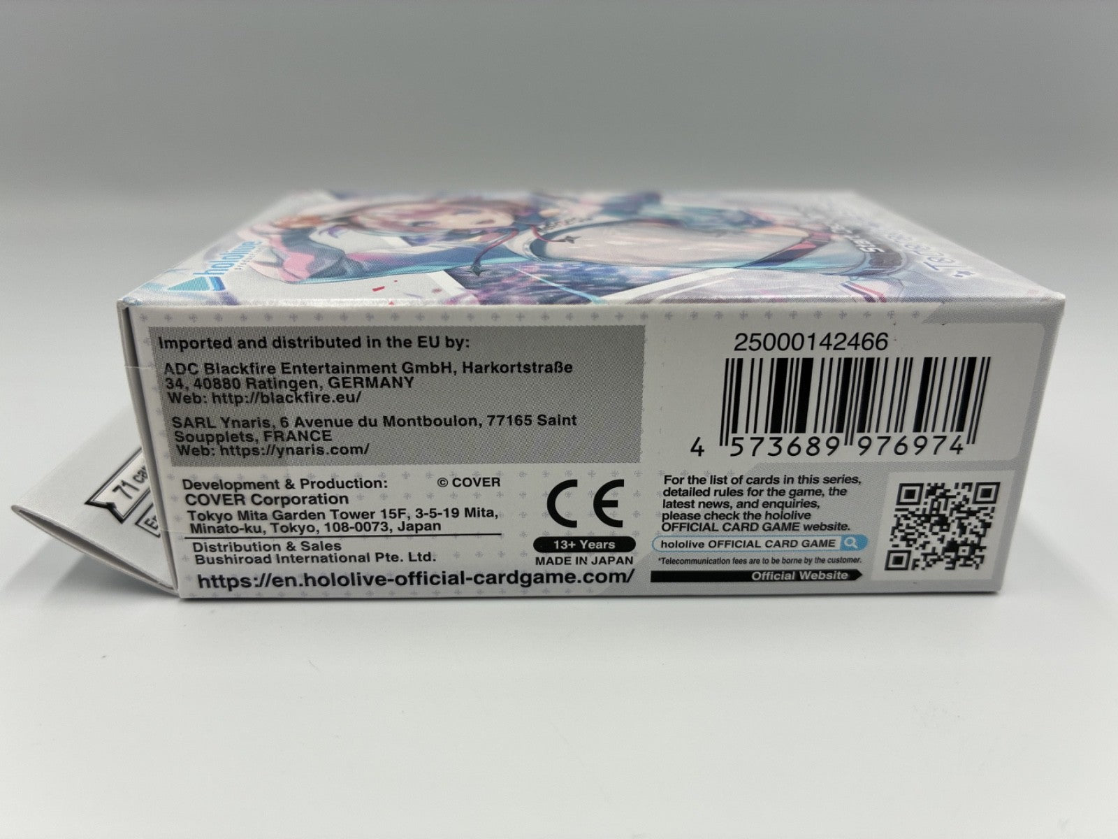 The back of a boxed card game package photographed on a light gray background. The box is mostly white with printed text, a barcode, a QR code, and anime-style character artwork across the top.